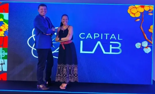Duas pessoas em pé ao lado do painel luminoso do Capital Lab